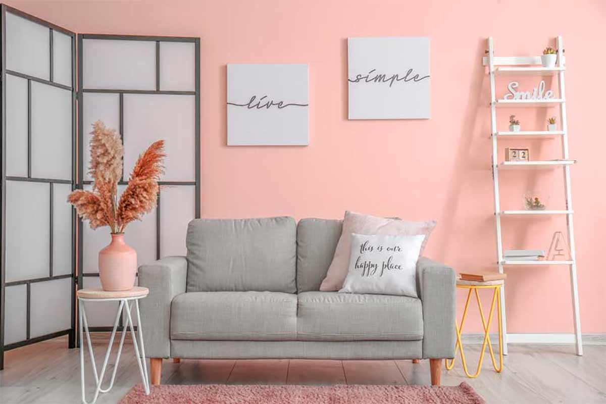 Salon avec canapé gris clair et mur rose avec des cadres blancs.